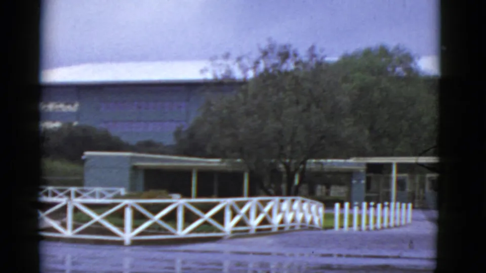 1952 California 8mm frame showing a modern flat-roof exterior, white fence, trees, and paved foreground.