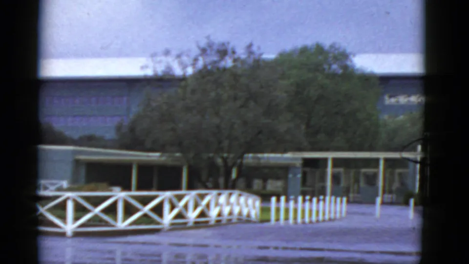 1952 California home-movie frame of a covered entry area, white fence, short pickets, and trees in front of a flat-roof building.