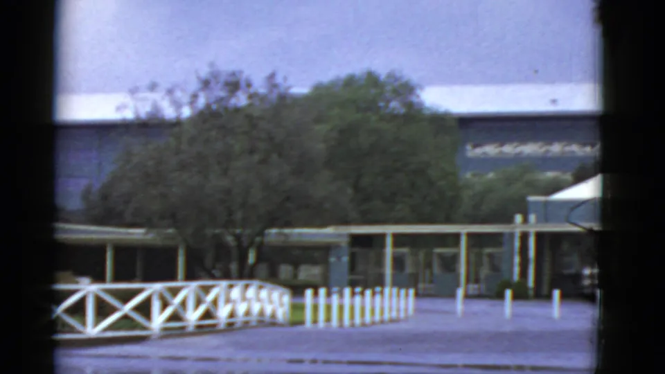 1952 California 8mm frame with white X-pattern fencing, short pickets, trees, and a low modern exterior in soft focus.