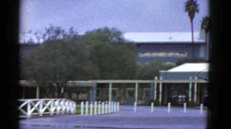 1952 California home-movie frame showing a low modern building, white fence, bollards, trees, and palms at the edge of the scene.