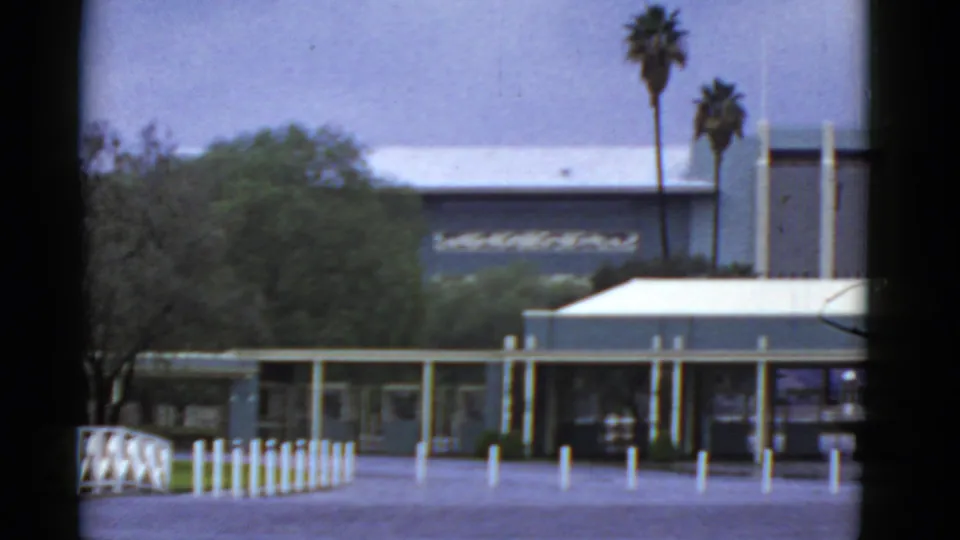 1952 California home-movie frame of a building entrance area with white bollards, a fence, trees, and palms.