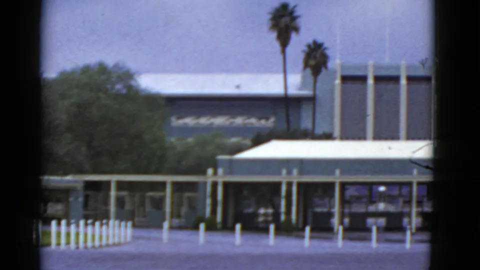 Closer 1952 California frame with a low building, covered walkway, white fence, bollards, trees, and palms.