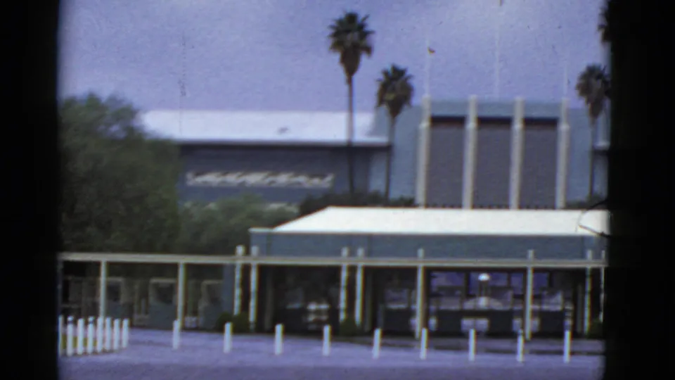 1952 California 8mm frame showing the building and covered entry framed by bollards, greenery, and palms.