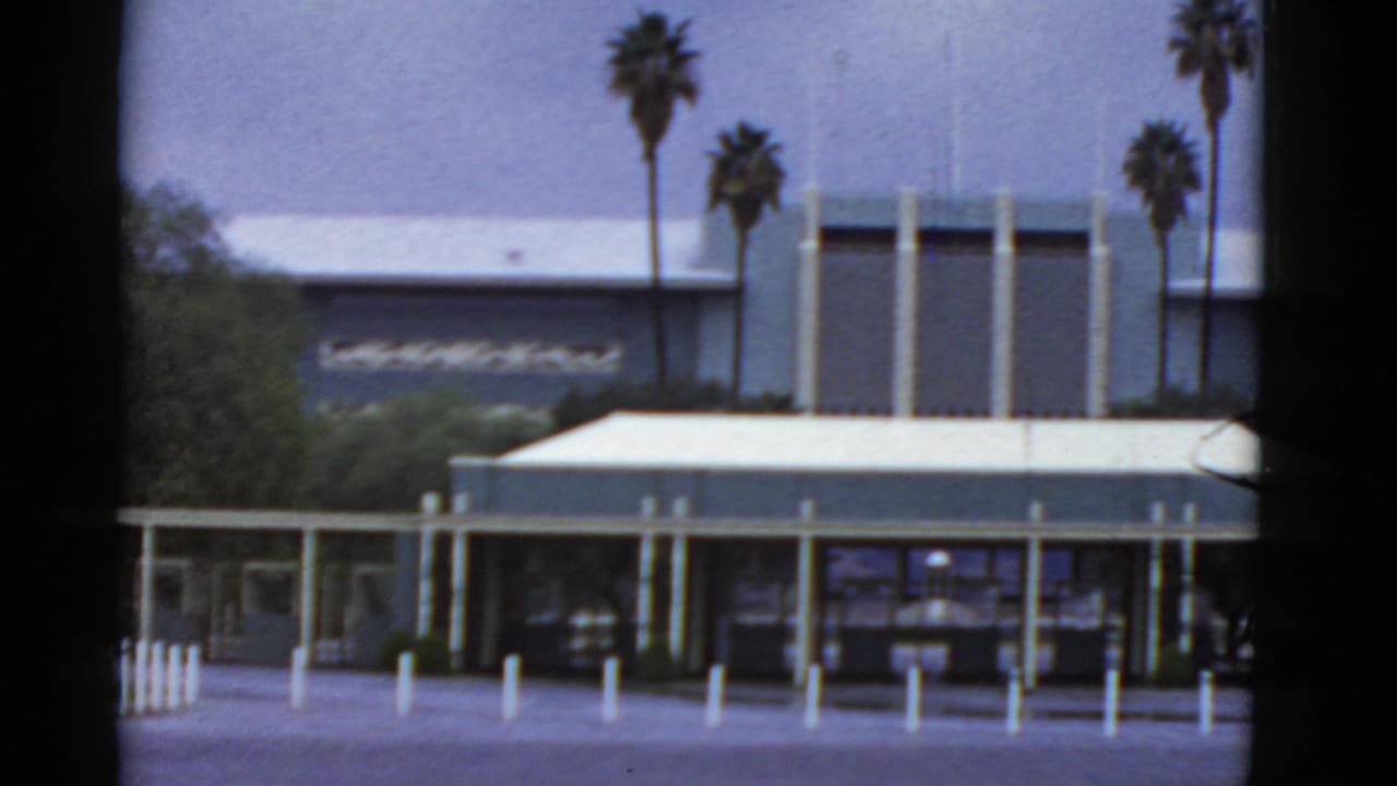 Distant flat-roofed modern building with palm trees and white bollards in front.