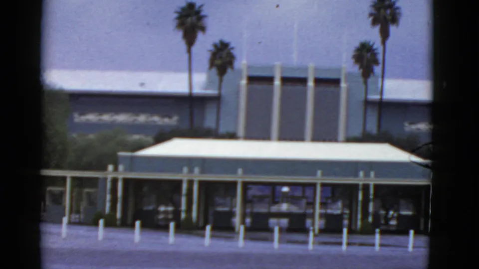 1952 California exterior view of a mid-century modern entrance with glass doors, white bollards, palms, and soft 8mm framing.