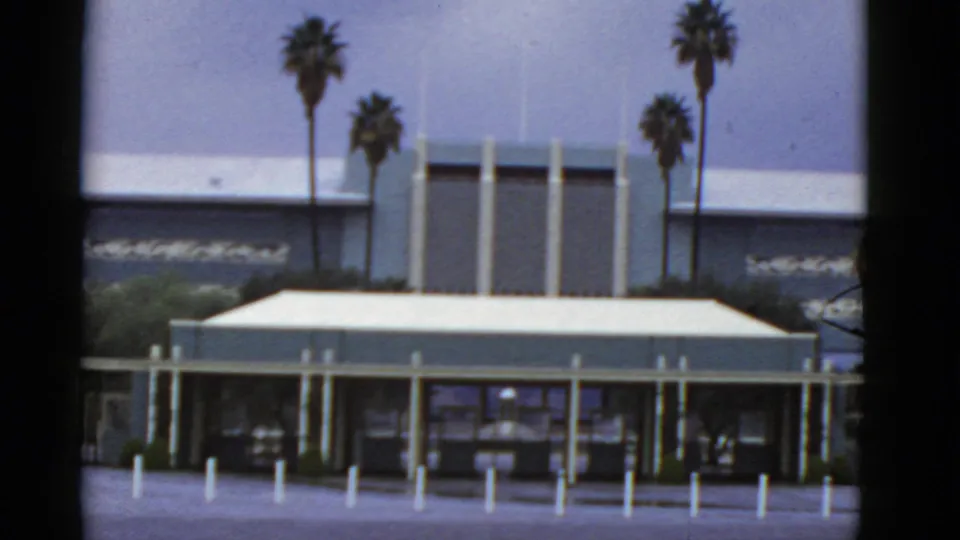 1952 California home-movie frame showing a closer view of the modern facade, vertical columns, flat roofline, palms, and foreground bollards.