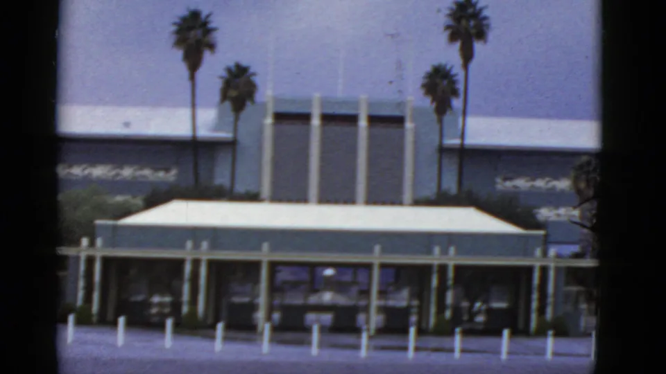 1952 California exterior frame of a low modern building with a flat canopy, glass frontage, bollards, palms, and faded blue sky.