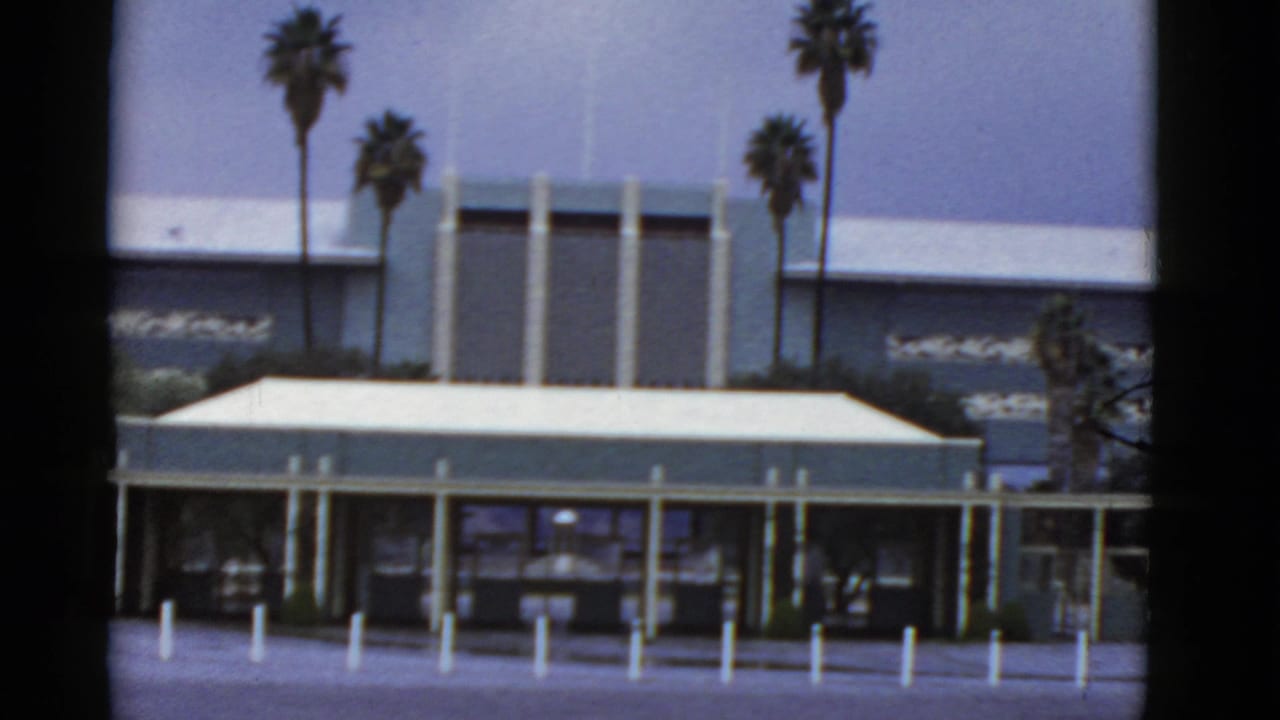 1952 California 8mm frame showing a low modernist building, vertical columns, palm trees, bollards, and an empty paved approach.