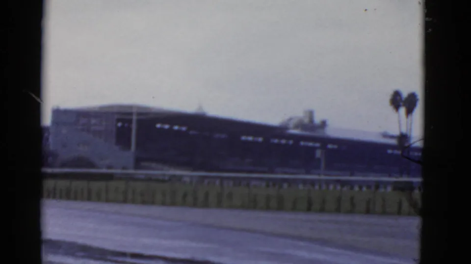 1952 California 8mm frame showing the side of a low modern building, a foreground barrier, broad pavement, and palm trees at right.