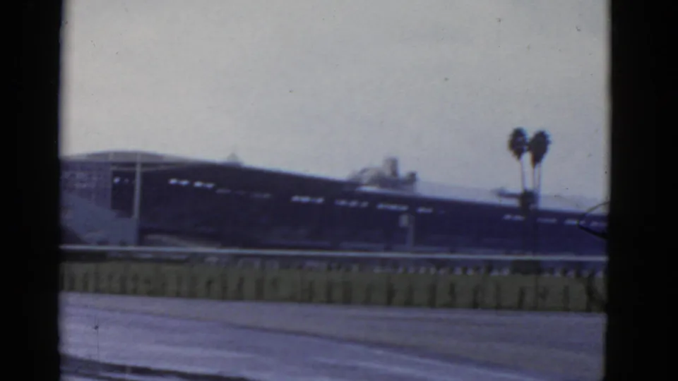 1952 California 8mm frame showing a low building across an empty paved area, with palm trees and a low barrier in the foreground.