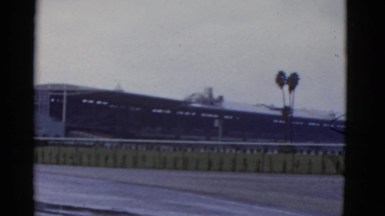 1952 California 8mm frame showing a blurry rail-side building, pale sky, paved foreground, and twin palm trees near the right edge.
