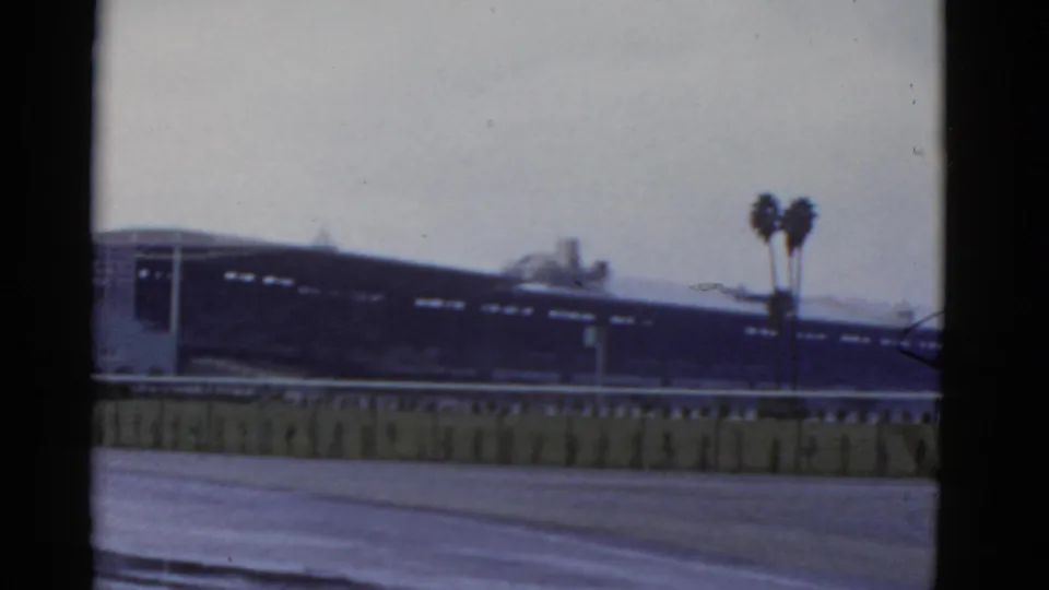 1952 California 8mm frame showing a blurry rail-side building, pale sky, paved foreground, and twin palm trees near the right edge.