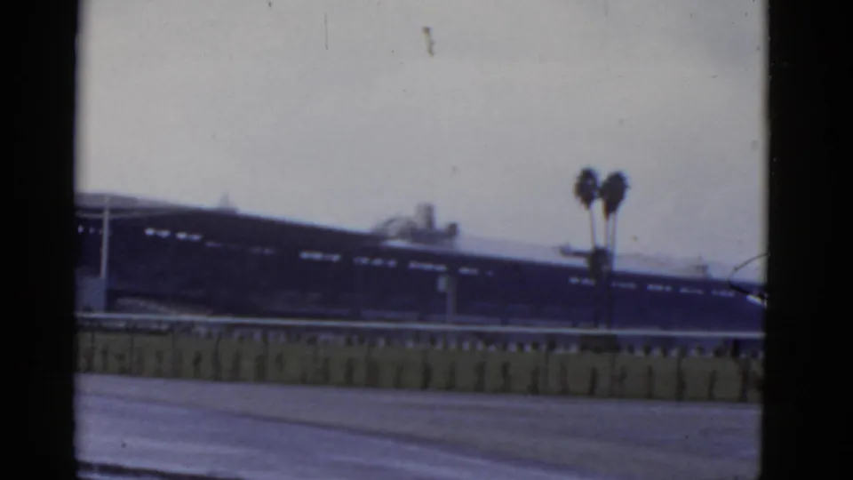 1952 California 8mm frame of a soft-focus exterior, likely rail-side, with a long dark structure, low barrier, and twin palm trees.