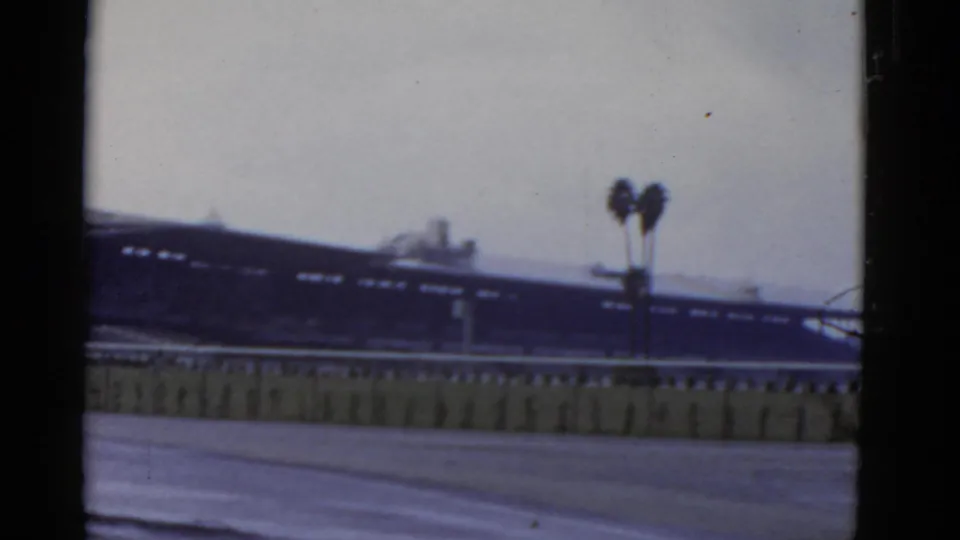 1952 California home-movie frame showing a blurred long building behind a low rail, with palm trees and visible 8mm frame edges.