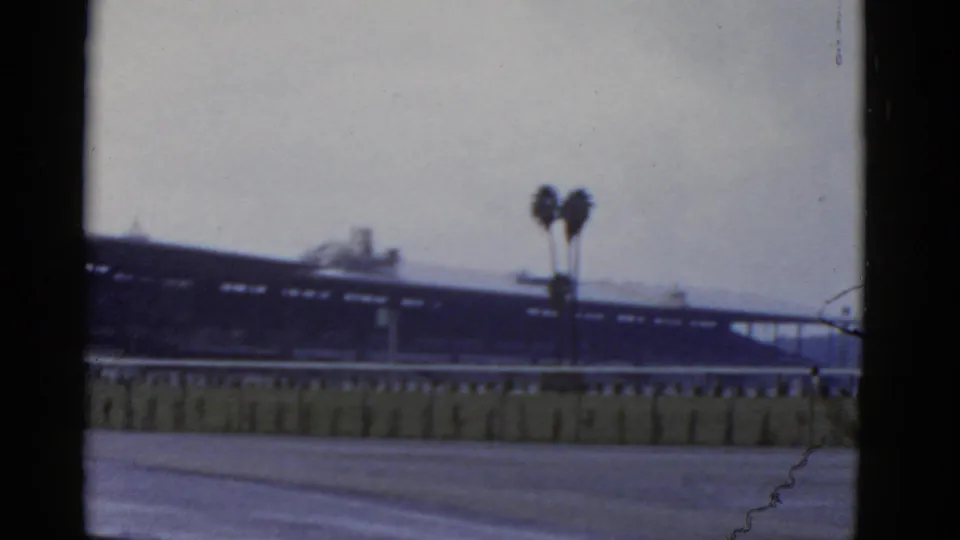 1952 California 8mm frame with a dark rail-side structure, pale sky, foreground paving, low barrier, and twin palm trees.