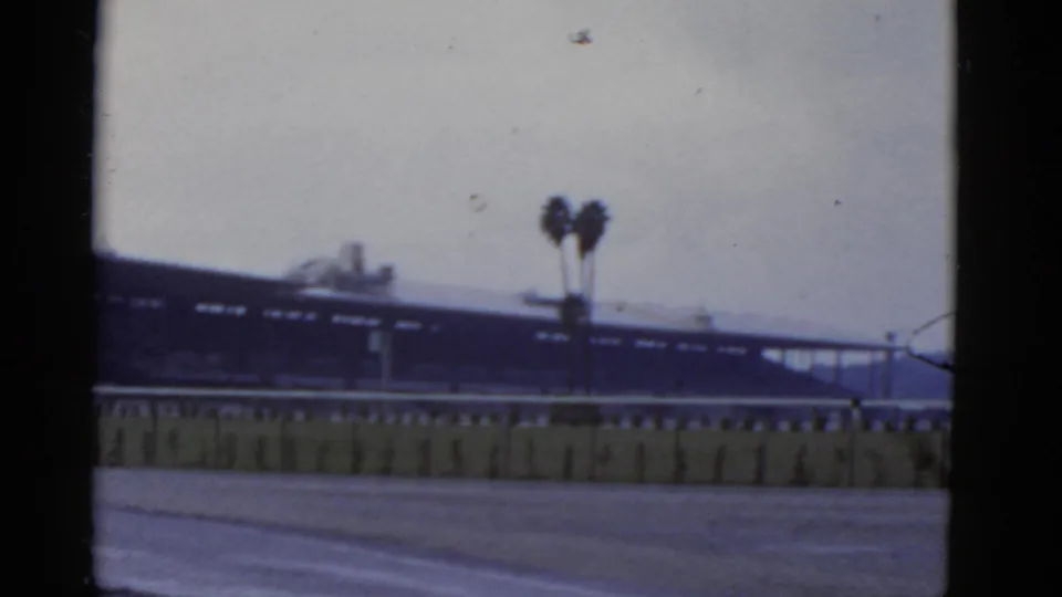 1952 California 8mm frame showing a blurred horizontal structure beyond a rail, two palm trees, open ground, and muted sky.