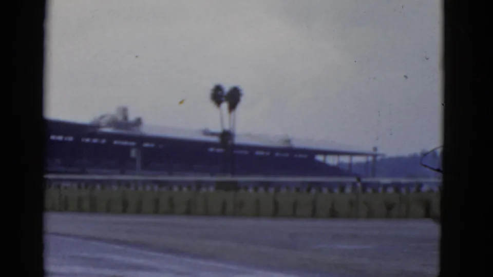 1952 California home-movie frame with a soft distant structure, rail or fence, paired palm trees, pale sky, and hillside at right.