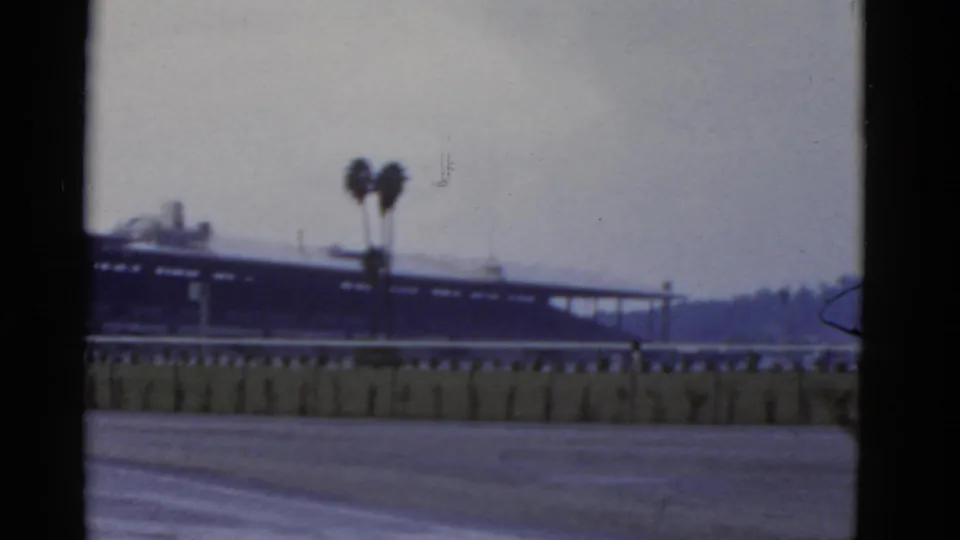 1952 California 8mm frame showing the structure partly shifted left, a rail in front, two palms, open ground, and visible film wear.