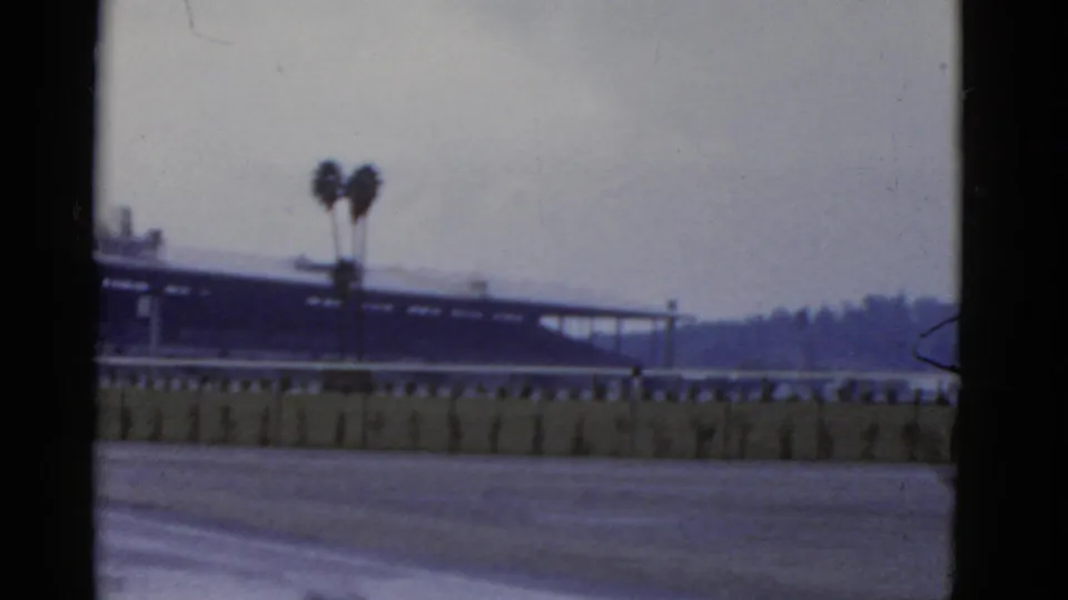 1952 California home-movie frame with a blurred structure behind a rail, two palm trees, open foreground, and distant hillside.