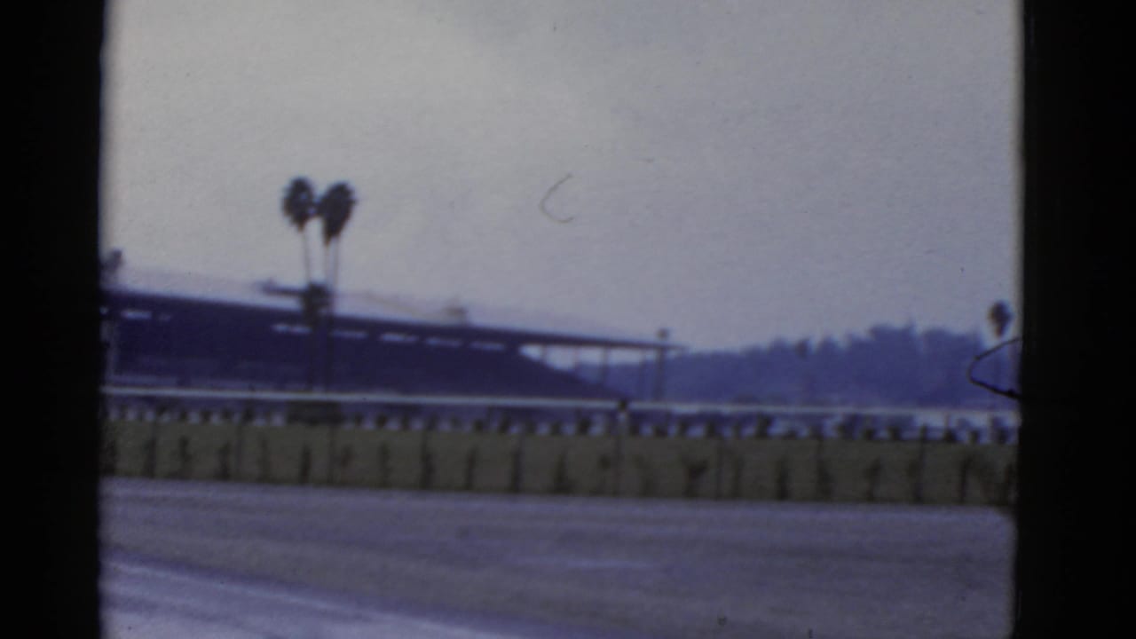 1952 California 8mm frame showing an empty racetrack edge, low barrier, distant grandstand, palms, and overcast sky.