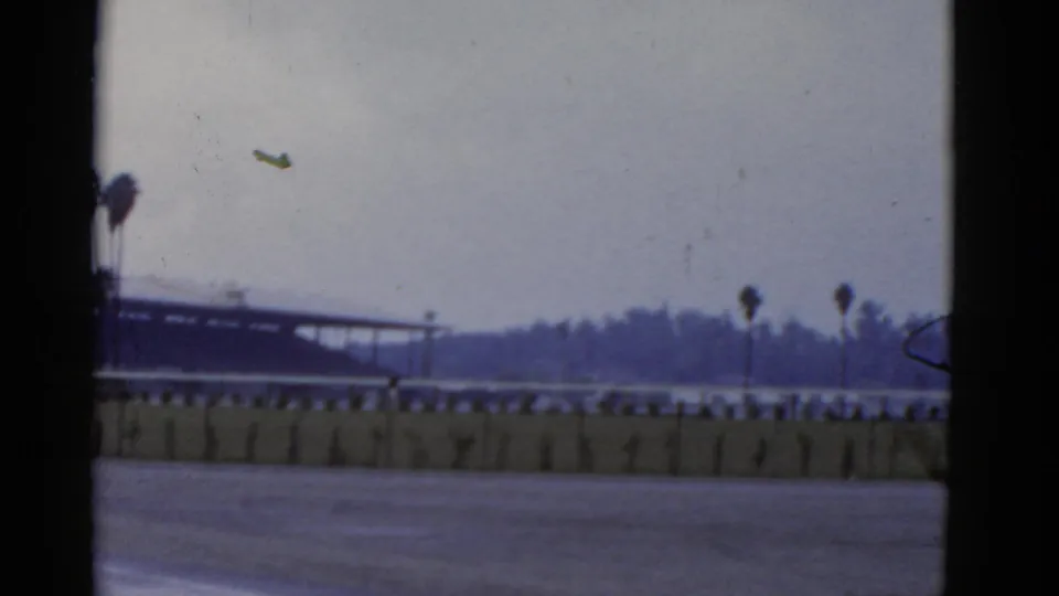 1952 California 8mm frame showing a small aircraft over an empty racetrack area with palms, low fence, and distant grandstand.