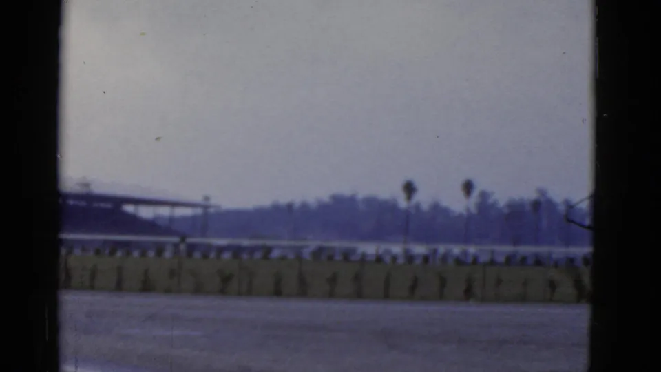 1952 California home-movie frame with an empty racetrack foreground, low barrier, palms, distant trees, and soft overcast light.