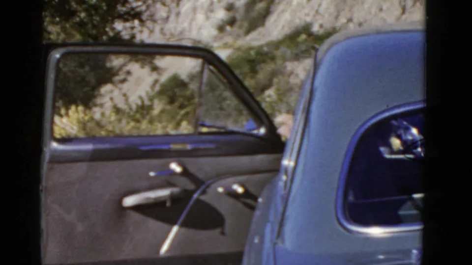 Open door of a 1950s car in a dry California mountain setting, with shrubs and rocky hillside behind it.
