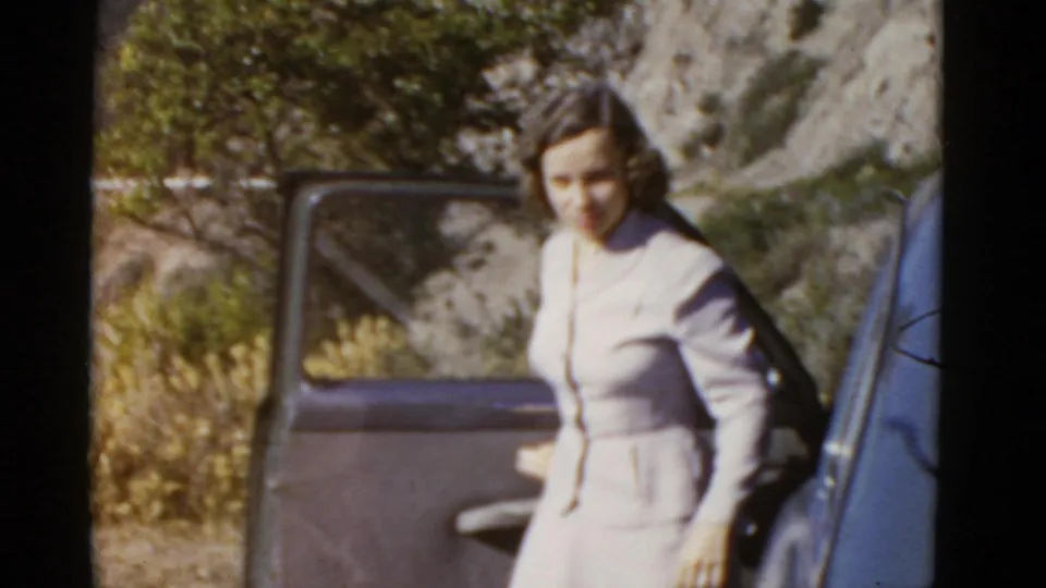 Adult woman in a light skirt suit stands beside an open 1950s car door in a rural California hillside setting.