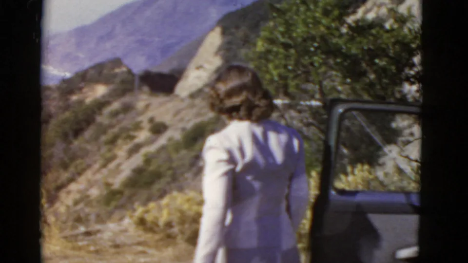 Woman viewed from behind near an open 1950s car door, with dry hills and mountains in the California landscape.