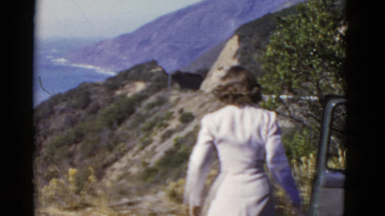 Woman in a pale dress seen from behind near a coastal hillside overlook.