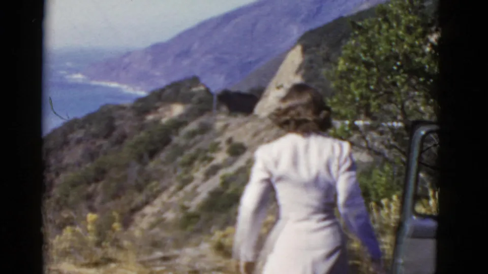 In 1952 California home-movie footage, a woman in a pale dress stands near brush and looks toward the Pacific coastline.
