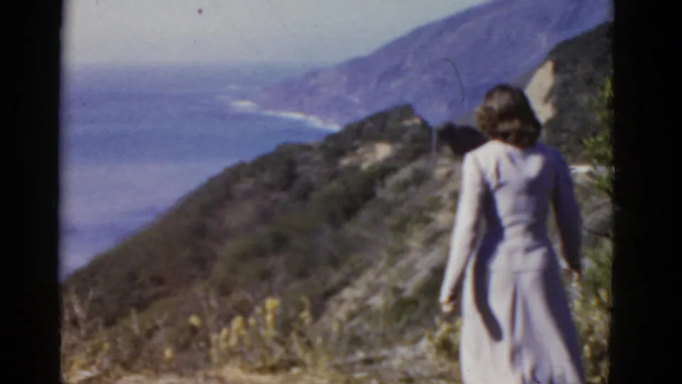 The woman moves along a California coastal slope, filmed from behind against the blue ocean and mountainous shoreline.