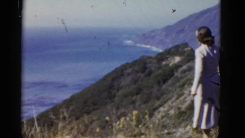 A woman in a light dress stands near the frame edge while the camera looks over California coastal hills and ocean water.