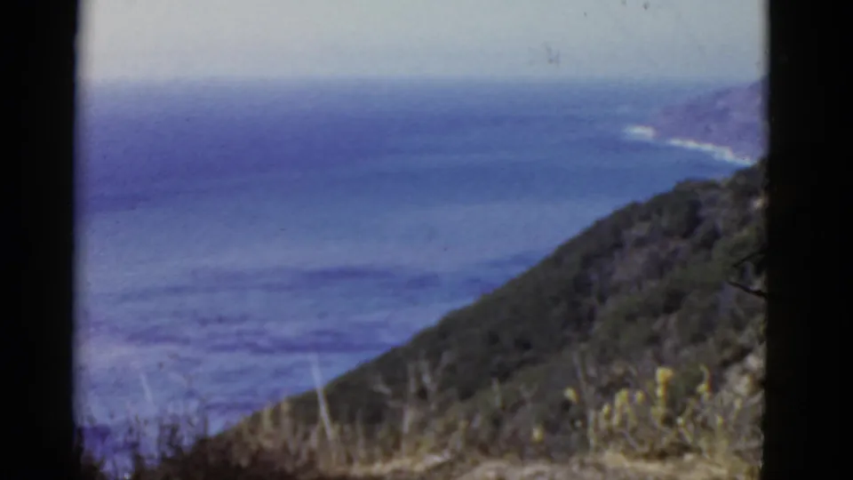 1952 California 8mm frame showing a green coastal slope above blue Pacific water, with brush and film borders visible.