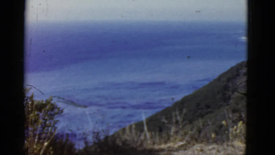 High 8mm view along the California coast, showing blue water, a sloping green headland, brush, and soft film texture.