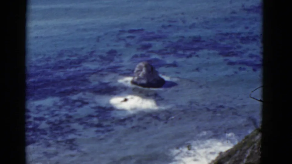 1952 California coastal frame with a rocky sea stack, pale breaking surf, dark kelp-like water patches, and a steep slope at the edge.