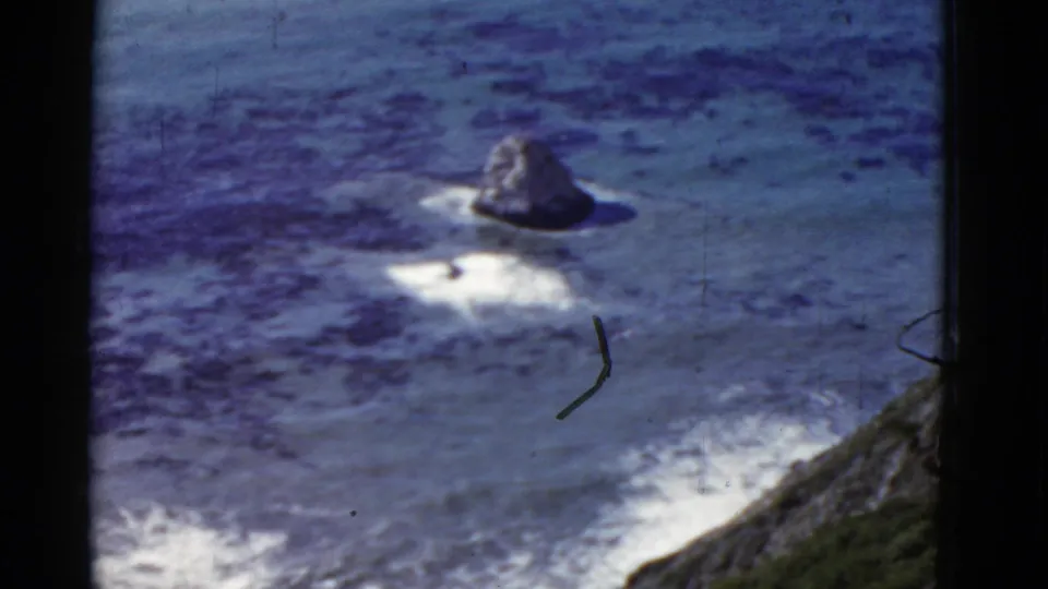 8mm California coastal view showing a sea stack, white surf, blue ocean, dark offshore patches, hillside edge, and visible film wear.