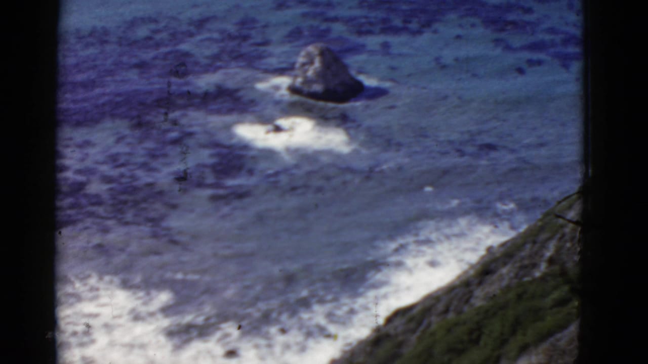1952 California 8mm frame showing a high vantage over blue ocean, white surf, an offshore rock, and a steep cliff edge.