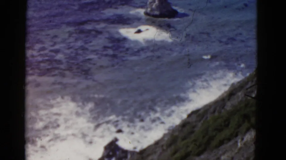 1952 California coastal frame with white surf, dark offshore rocks, and a sloping cliffside seen from above.