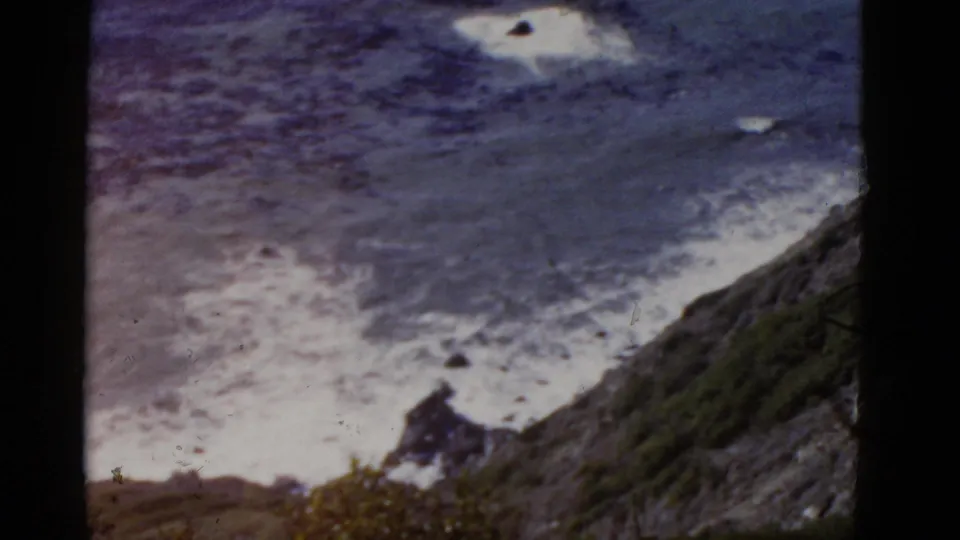 High-angle 1952 California 8mm view of surf, shoreline rocks, and a steep vegetated cliff descending toward the water.