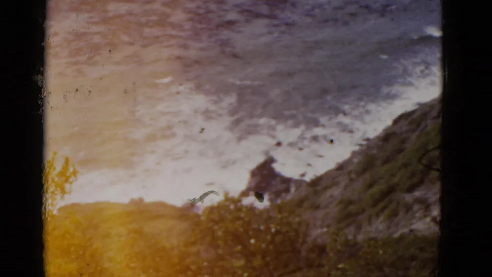 1952 California 8mm frame showing surf and a rocky cliffside with visible color fading and warm light leak near the lower edge.