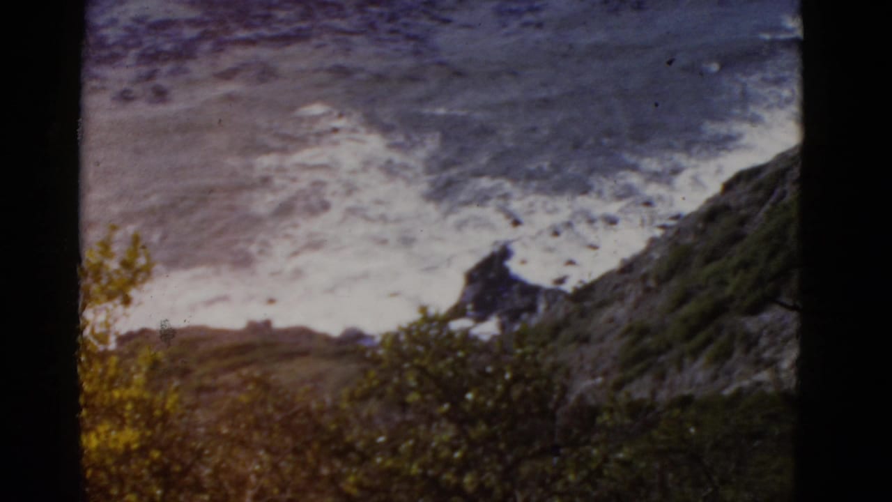 1952 California coastal home-movie frame with white surf hitting rocky shoreline beyond hillside foliage and dark gate edges.