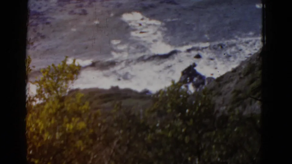 1952 California coast view with surf washing over rocks, scrubby yellow-green foliage, and heavy dark film-frame borders.