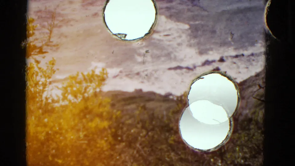 1952 California shoreline frame showing waves, brushy hillside, dark gate edge, and several bright circular damage spots on the film.