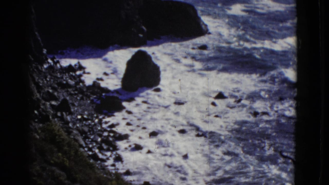 Foamy surf moves around rocks below a dark coastal cliff.