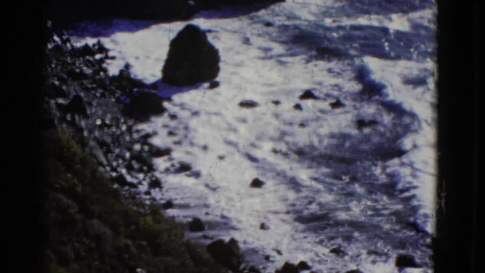 1952 8mm San Francisco shoreline view with waves breaking around scattered rocks and a dark sea stack near the surf line.