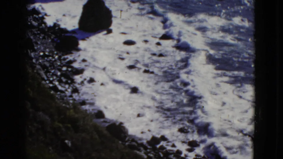 1952 8mm San Francisco coastal frame showing tide foam spreading over wet sand or rock, with dark shoreline vegetation at the edge.