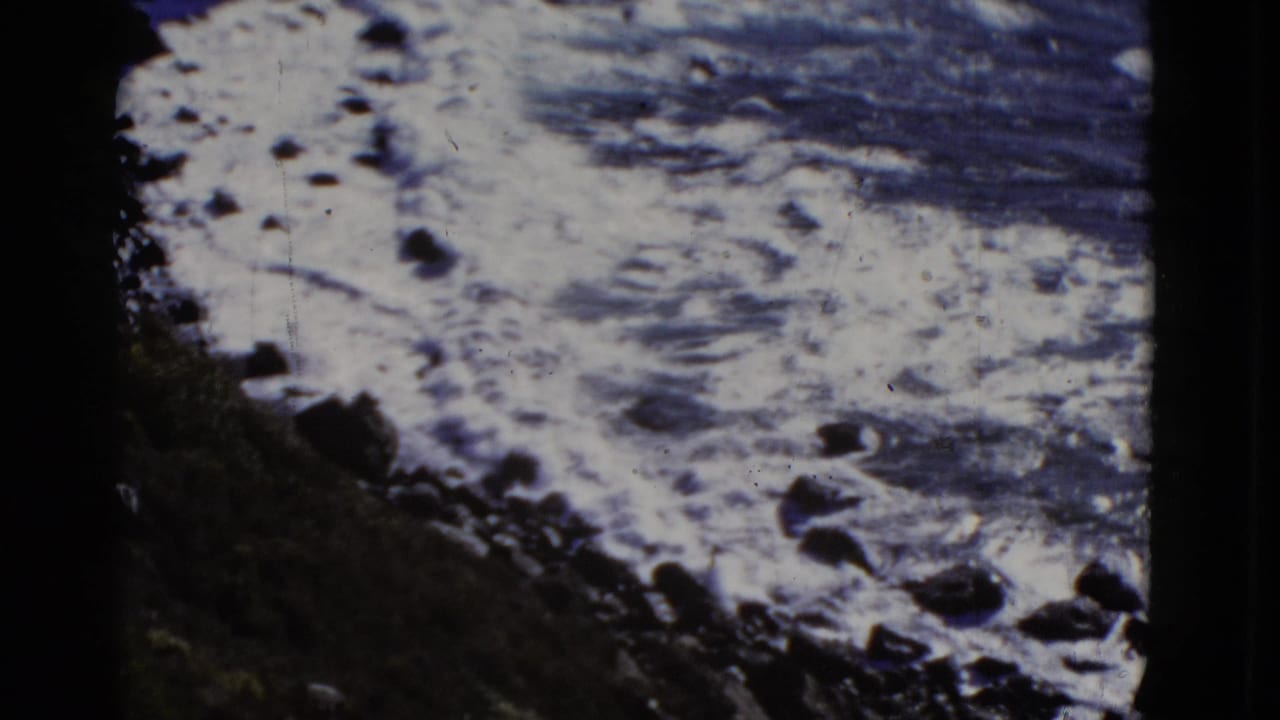 Foaming ocean water washes against dark rocks below a shadowed rocky slope.