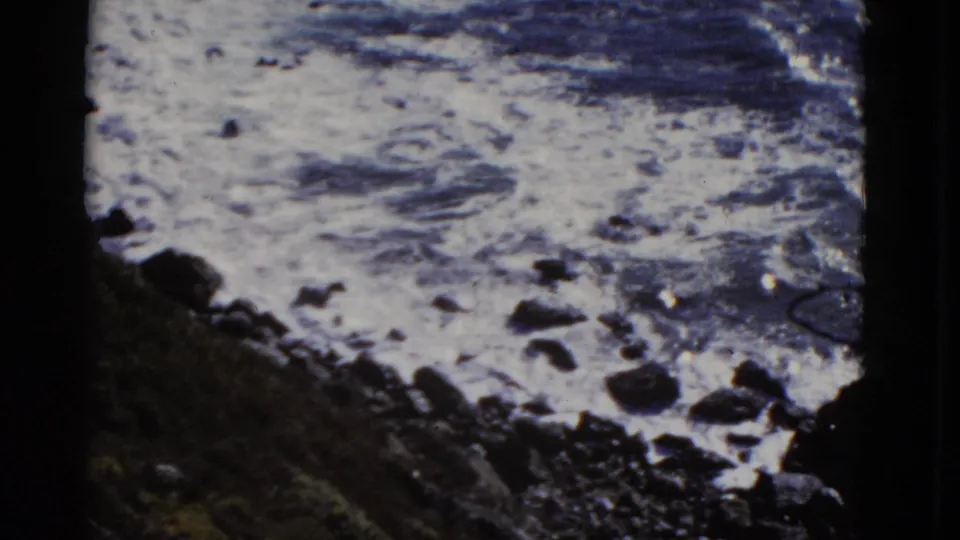 1952 San Francisco coastal frame with churning pale surf, scattered dark stones, and a sloped rocky shoreline in shadow.