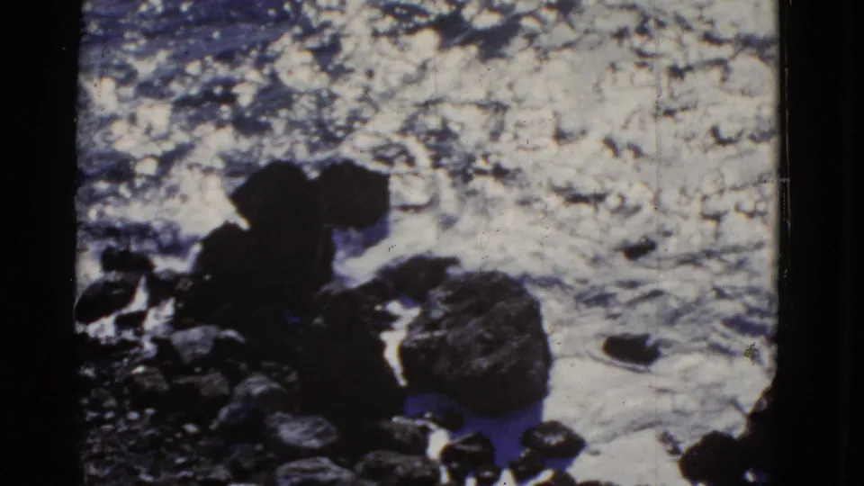 1952 San Francisco 8mm frame showing large foreground rocks silhouetted against bright, foaming ocean water.